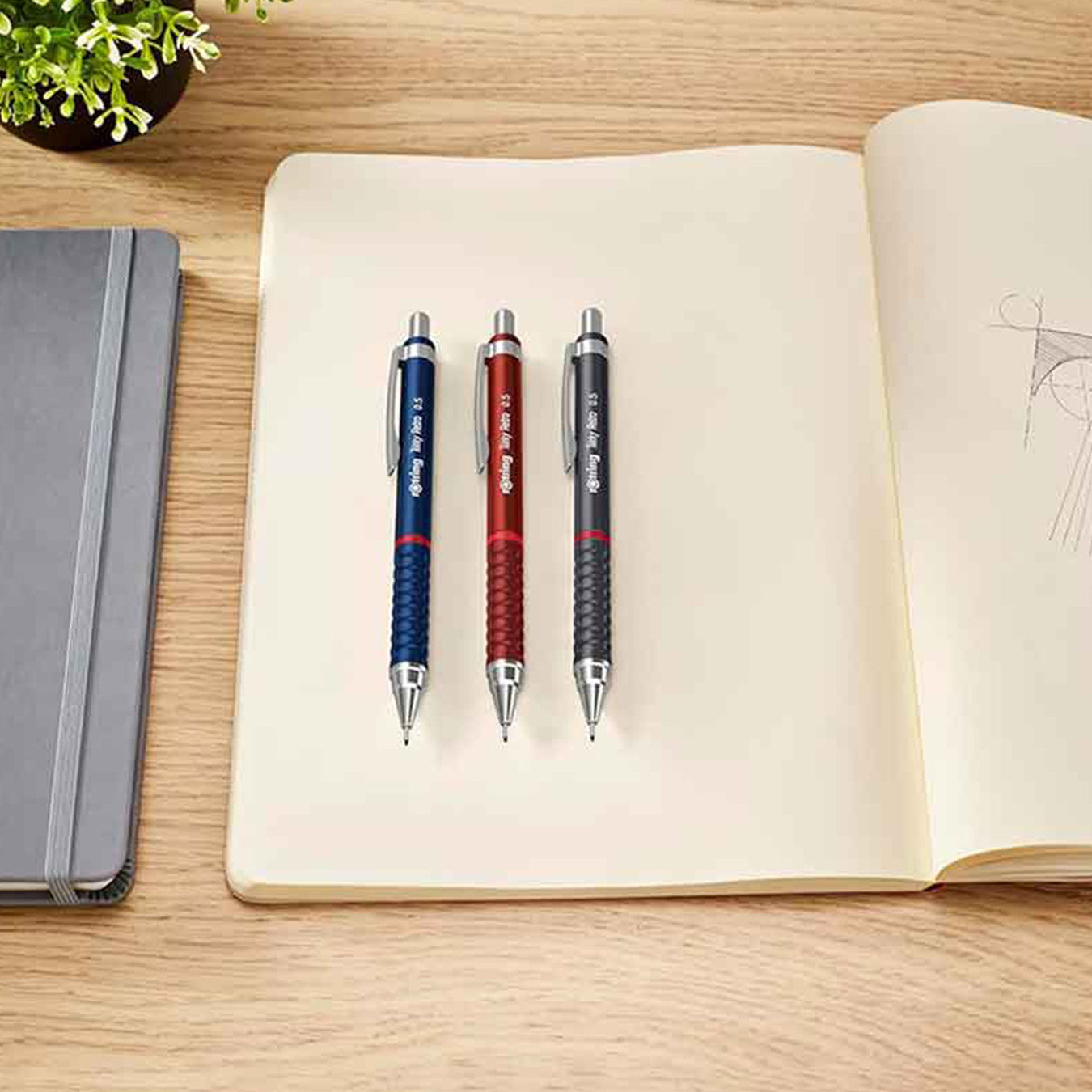 3 Rotring Tikky Retro 0.7mm Mechanical Pencil in different colours on an open notebook with a wooden surface and plant in the background